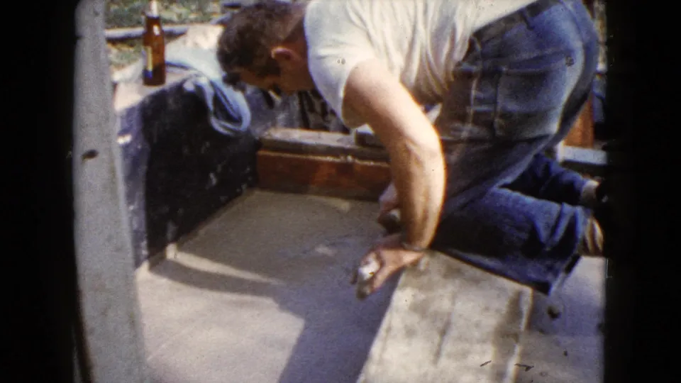 The 8mm frame shows a kneeling man working a trowel over fresh cement on a porch or patio in Wauconda.