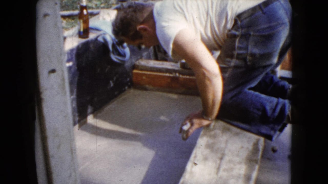 In 1960 Wauconda home-movie footage, a man in work clothes leans over wet pavement near a doorway and wooden form.