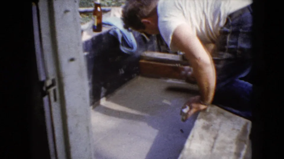 The man continues finishing the pavement surface near a wooden form, with a brown bottle and dark cloth behind him.