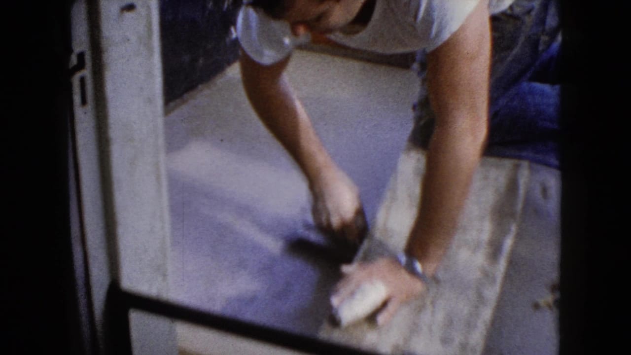 In supported 1960 Wauconda, Illinois footage, a man works close to a wet concrete step with a trowel while framed by a doorway or railing.