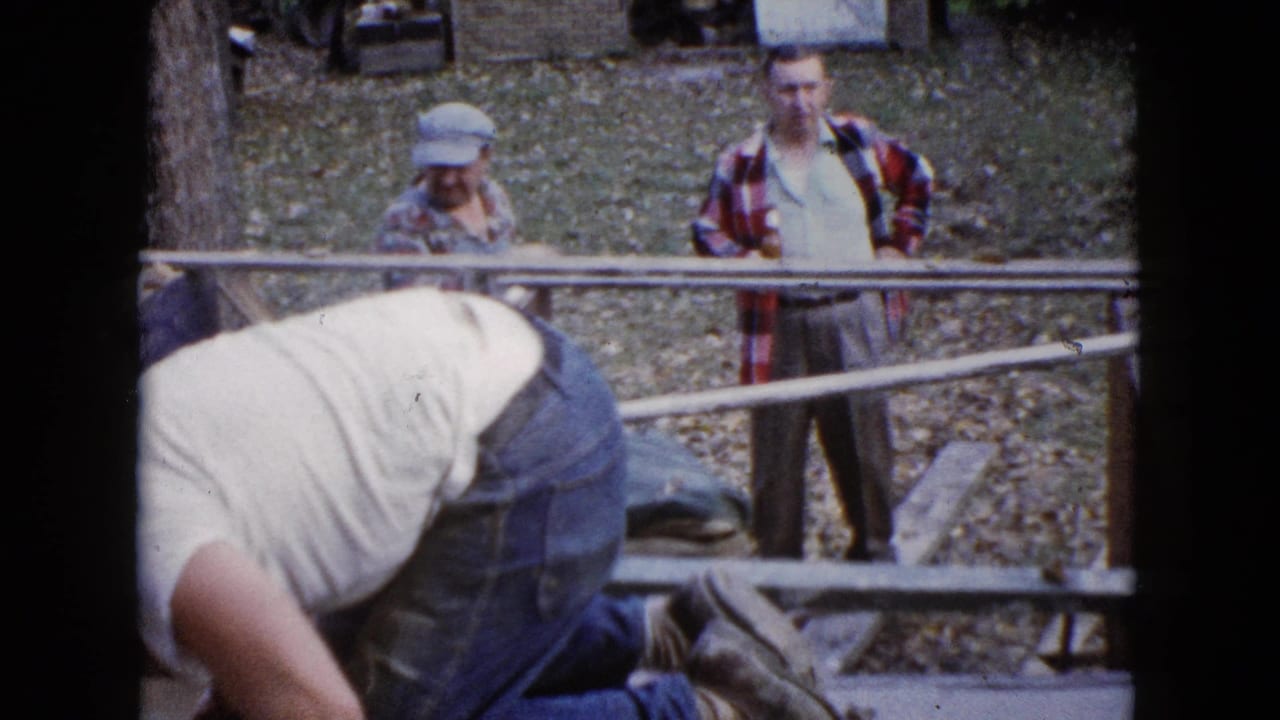 In the outdoor residential scene, two adult men observe from behind metal railings as the worker bends over the concrete step in the foreground.