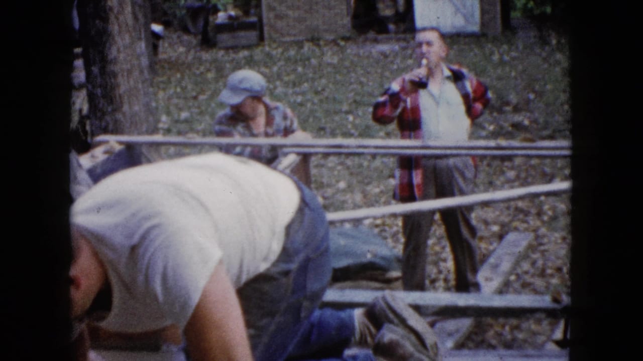 1960 Wauconda, Illinois home-movie frame showing men near a wood structure, one kneeling at work while another drinks from a bottle.
