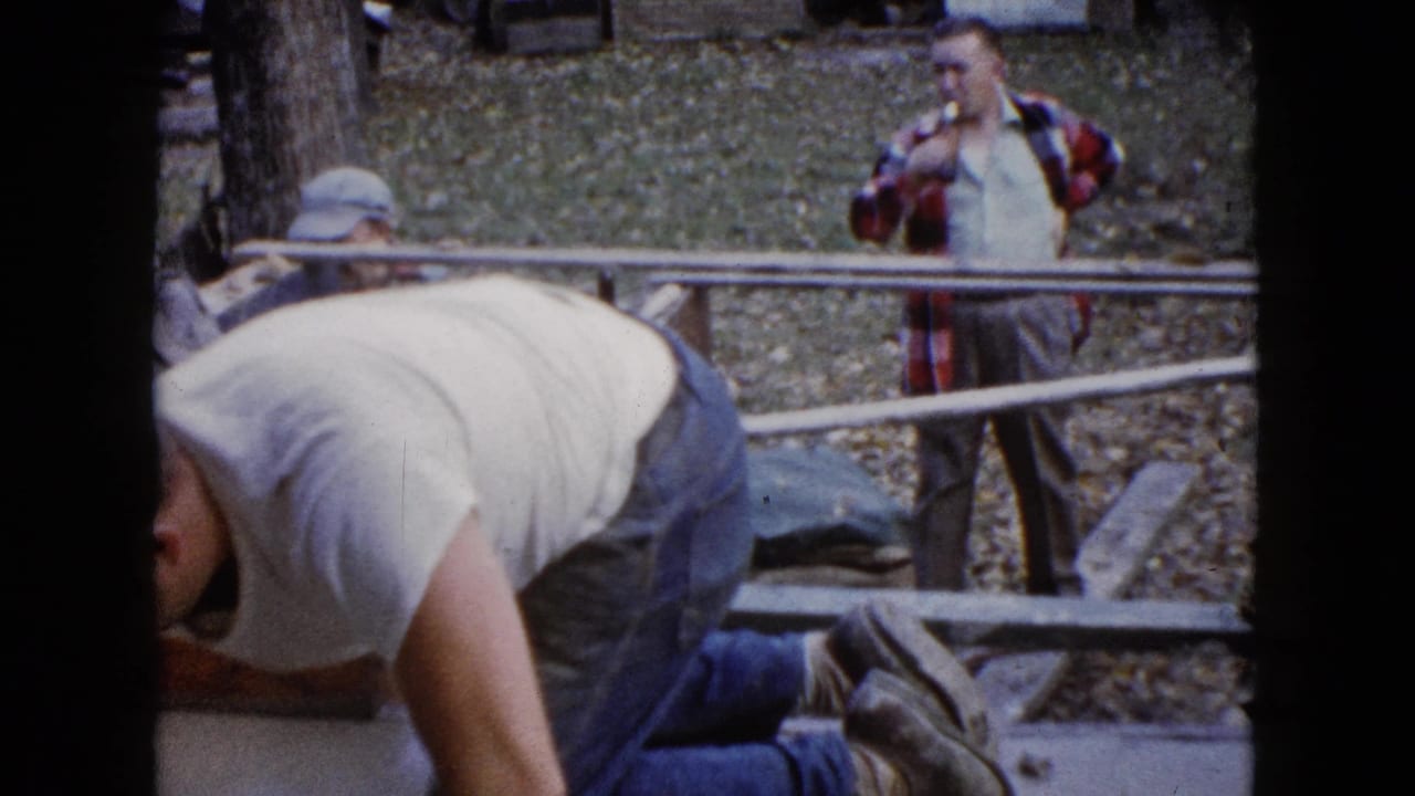 A kneeling man works in the foreground while two men stand behind wood rails in a leafy yard.