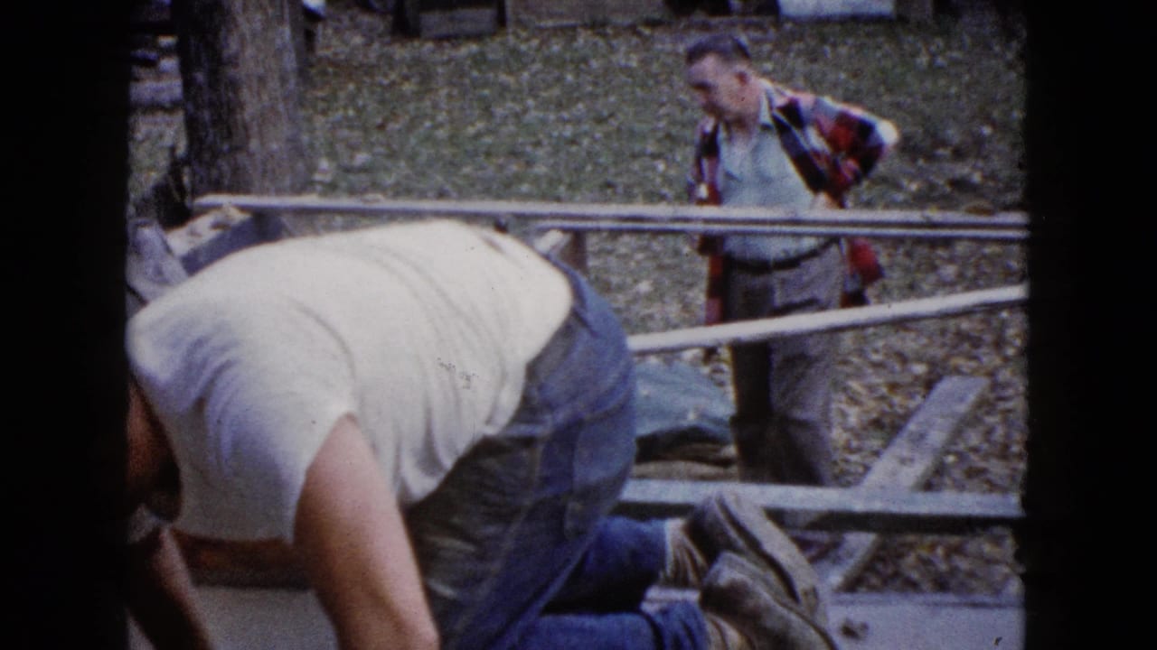 Home-movie frame from 1960 Wauconda showing a man bent over outdoor work while another adult in plaid stands in the yard.