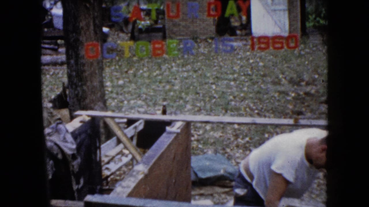 Man in a white shirt bends beside a wooden structure under colorful date letters.