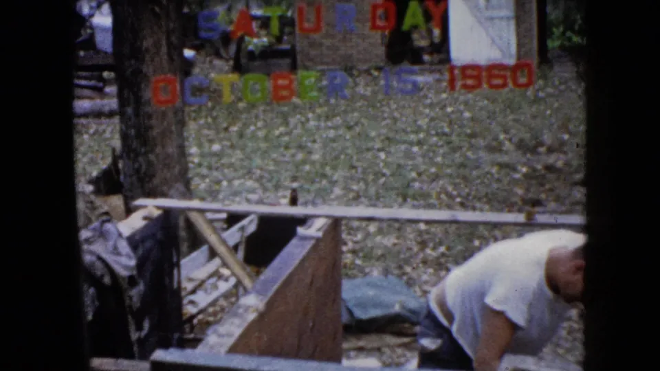 1960 Wauconda home-movie frame showing a man bending near lumber and a wooden structure with Saturday October 15 1960 lettering behind him.