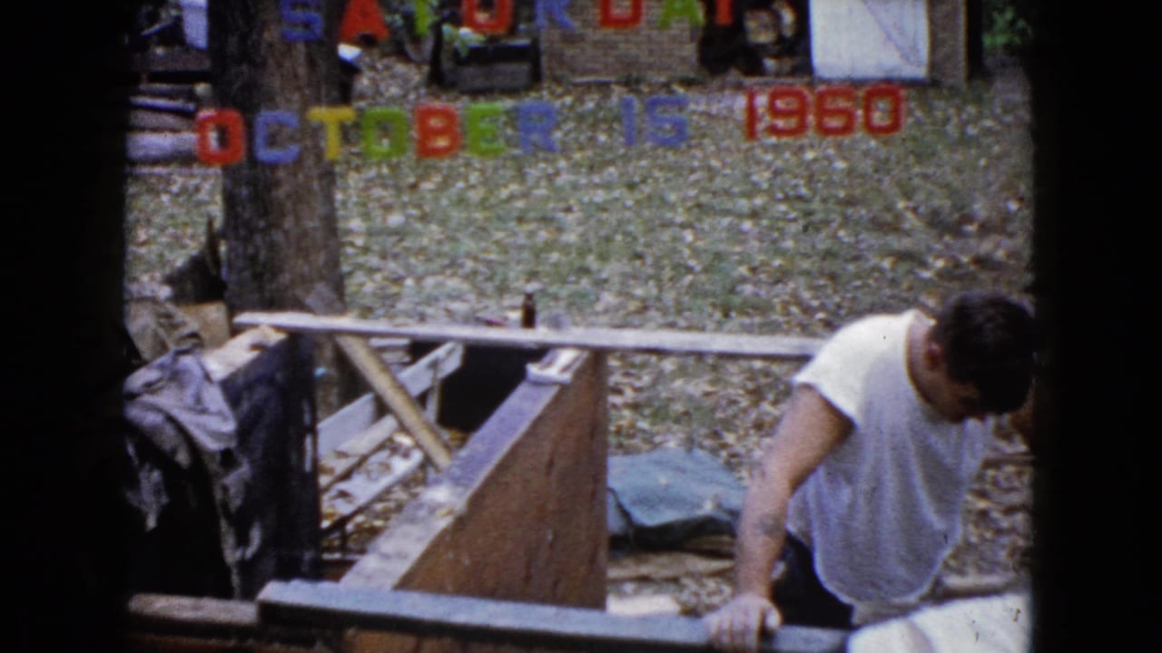 1960 Wauconda frame showing a man in a white shirt rising beside a wooden structure, with another partially visible person near the date letters.