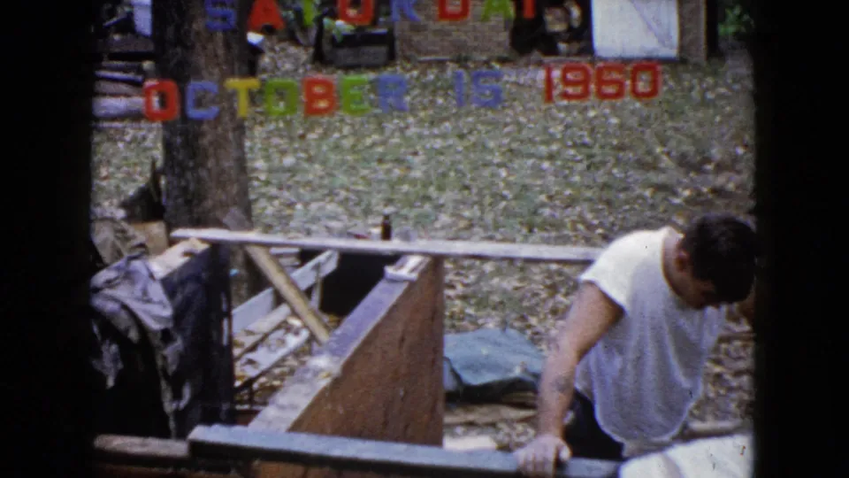 1960 Wauconda frame showing a man in a white shirt rising beside a wooden structure, with another partially visible person near the date letters.