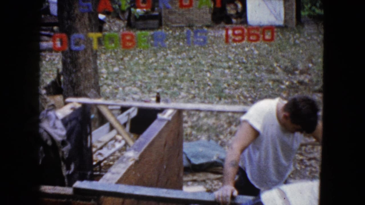 Man in a white sleeveless shirt works beside wooden boards in a leafy backyard.