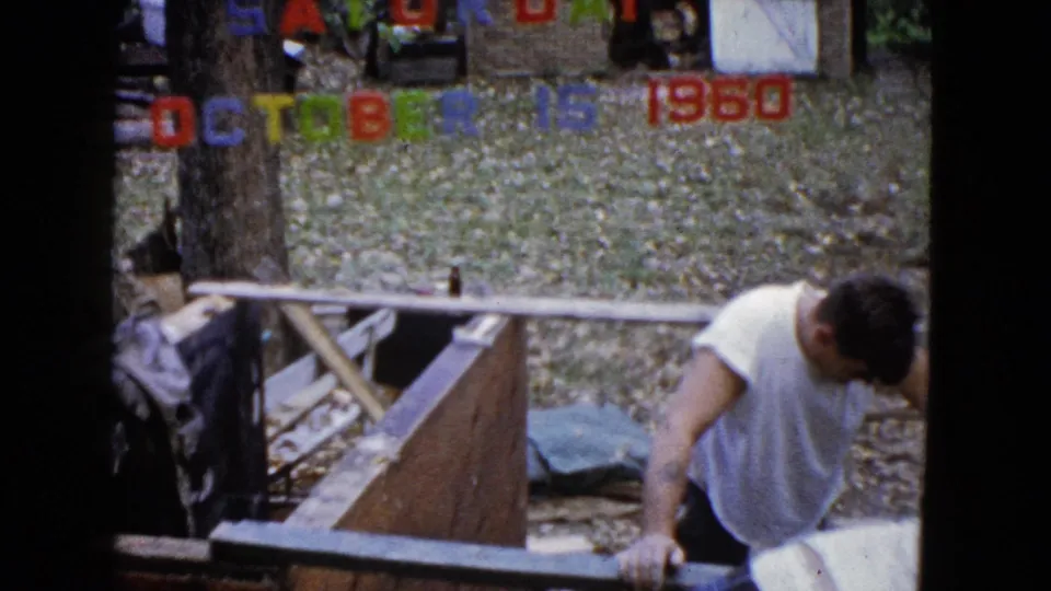 In Wauconda, Illinois in 1960, a man in a white sleeveless shirt works near a wooden structure beneath colorful date lettering.