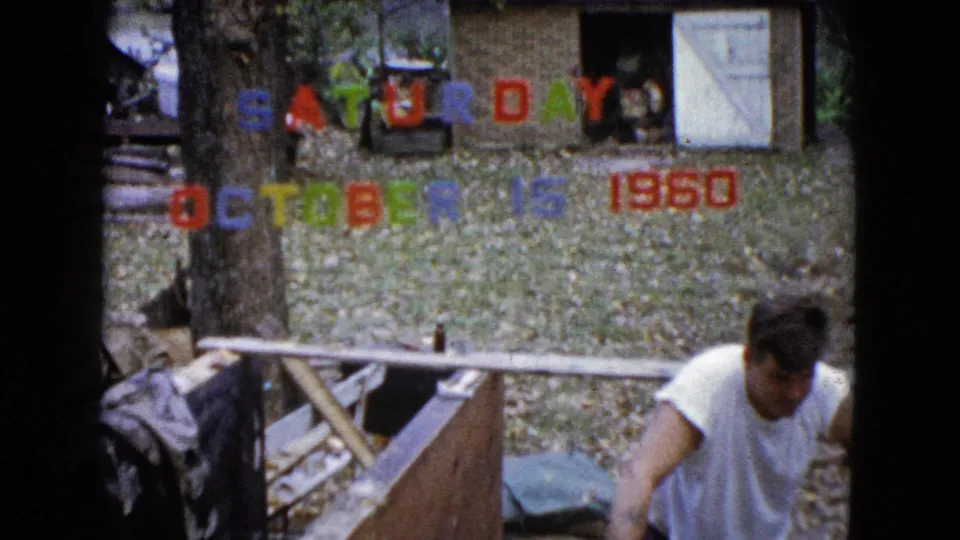 The 1960 home-movie frame shows the man bending at the work area in a Wauconda yard, surrounded by leaves and colored date lettering.