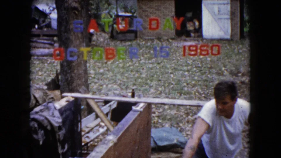 At the end of the 1960 Wauconda clip, the man remains beside the wooden work area with trees, leaves, and date letters in view.