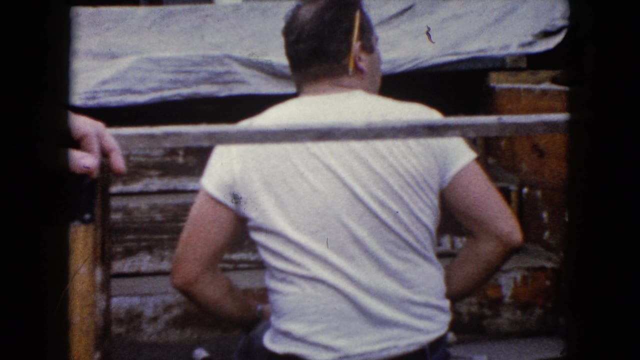 Man in a white T-shirt seen from behind beside rough wooden boards.