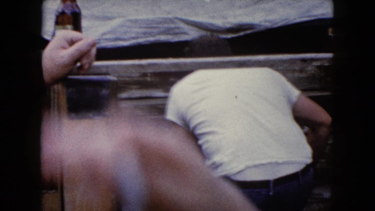 1960 Wauconda, Illinois home-movie frame with a bent-over worker and a nearby hand holding a glass bottle in the foreground.