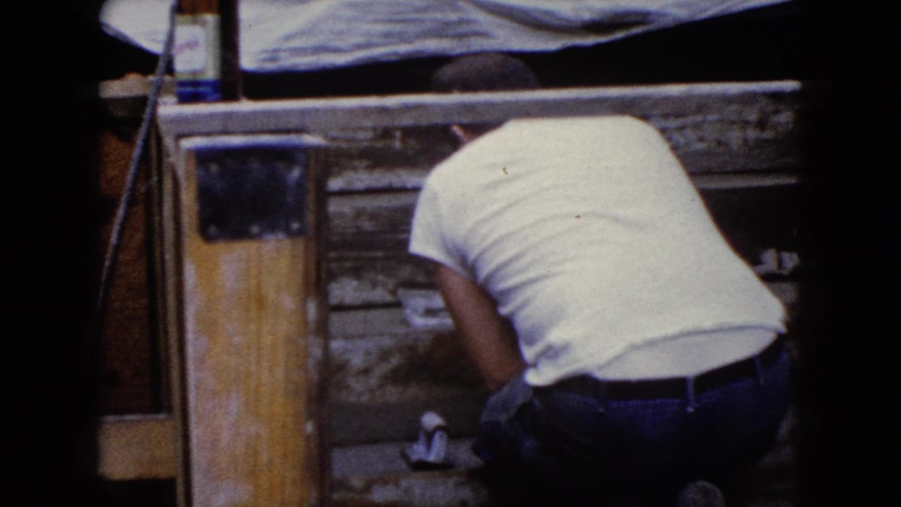 1960 Wauconda, Illinois 8mm frame showing a man in jeans bending at a low work area beside weathered wood.