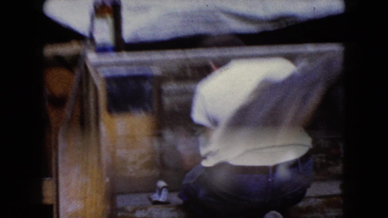 Soft 1960 Wauconda, Illinois 8mm frame of a man bending near a wooden structure, with a bottle visible near the left edge.
