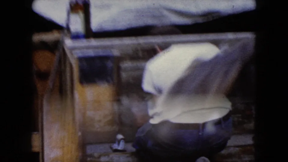 Soft 1960 Wauconda, Illinois 8mm frame of a man bending near a wooden structure, with a bottle visible near the left edge.