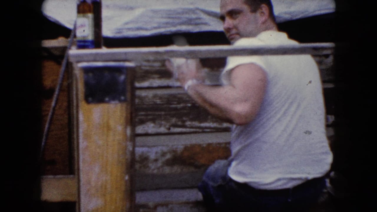 Man in a white T-shirt sanding or cleaning weathered wood beside a glass bottle.