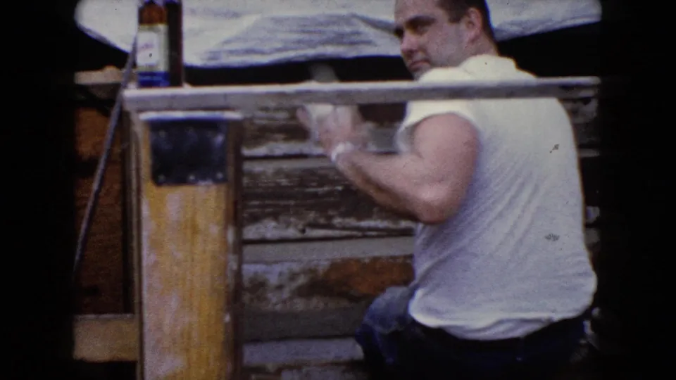 In Wauconda, Illinois in 1960, a man in a white T-shirt works on a weathered wooden structure with a glass bottle nearby.
