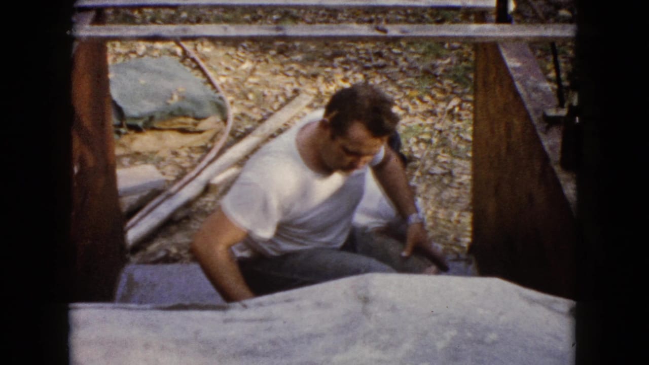 The camera looks down as the man crouches near the project area, surrounded by gravel, lumber, and a pale covered surface.