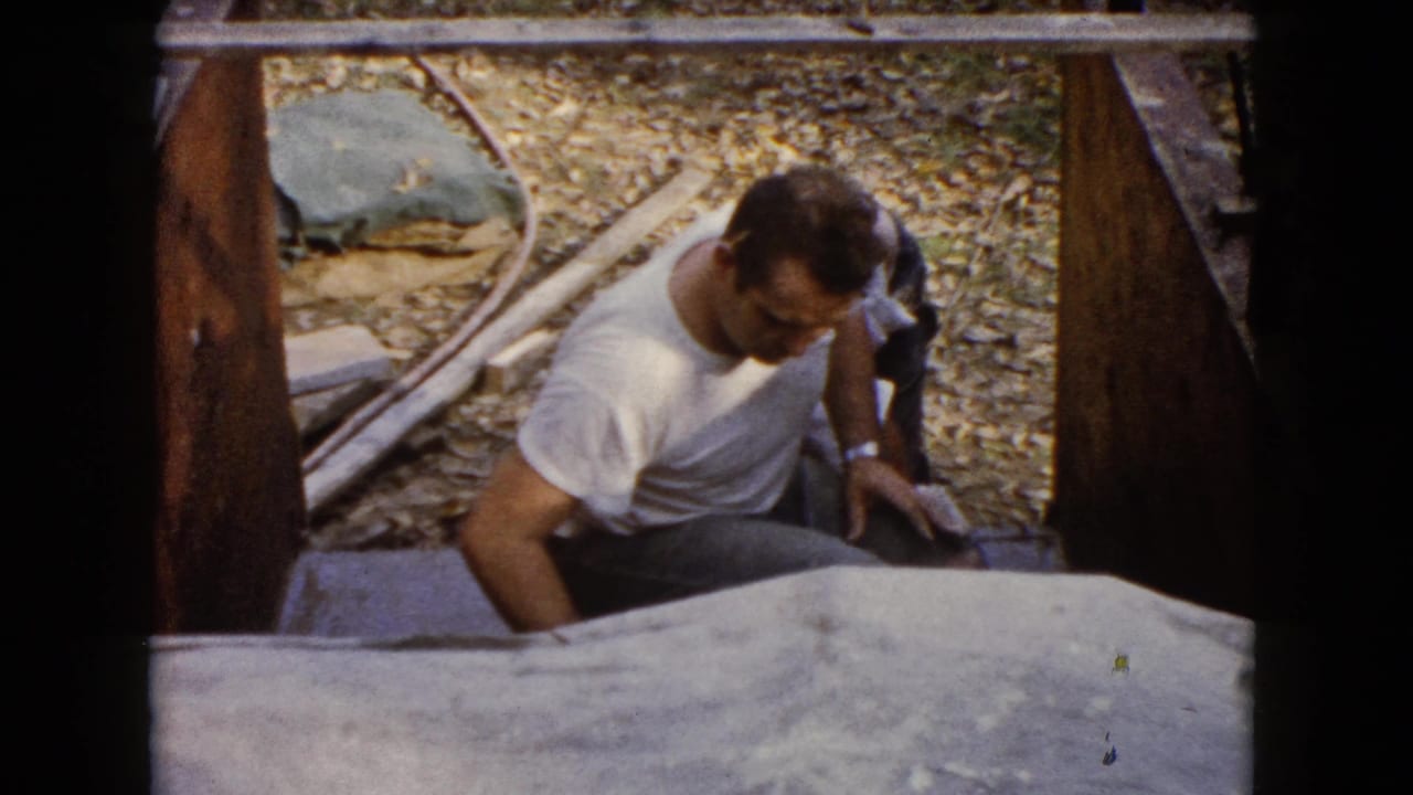 The man remains crouched beside the wooden structure in the Wauconda work area, with gravel, lumber, and soft 8mm color visible.