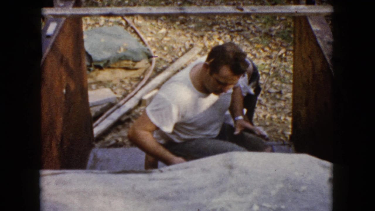 Man in a white T-shirt works beside gray concrete within wooden framing.