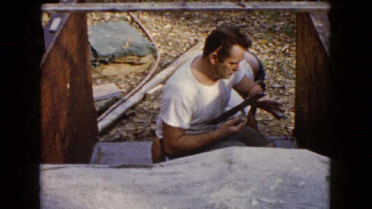Man holds a hand tool while seated or crouched beside the concrete form.