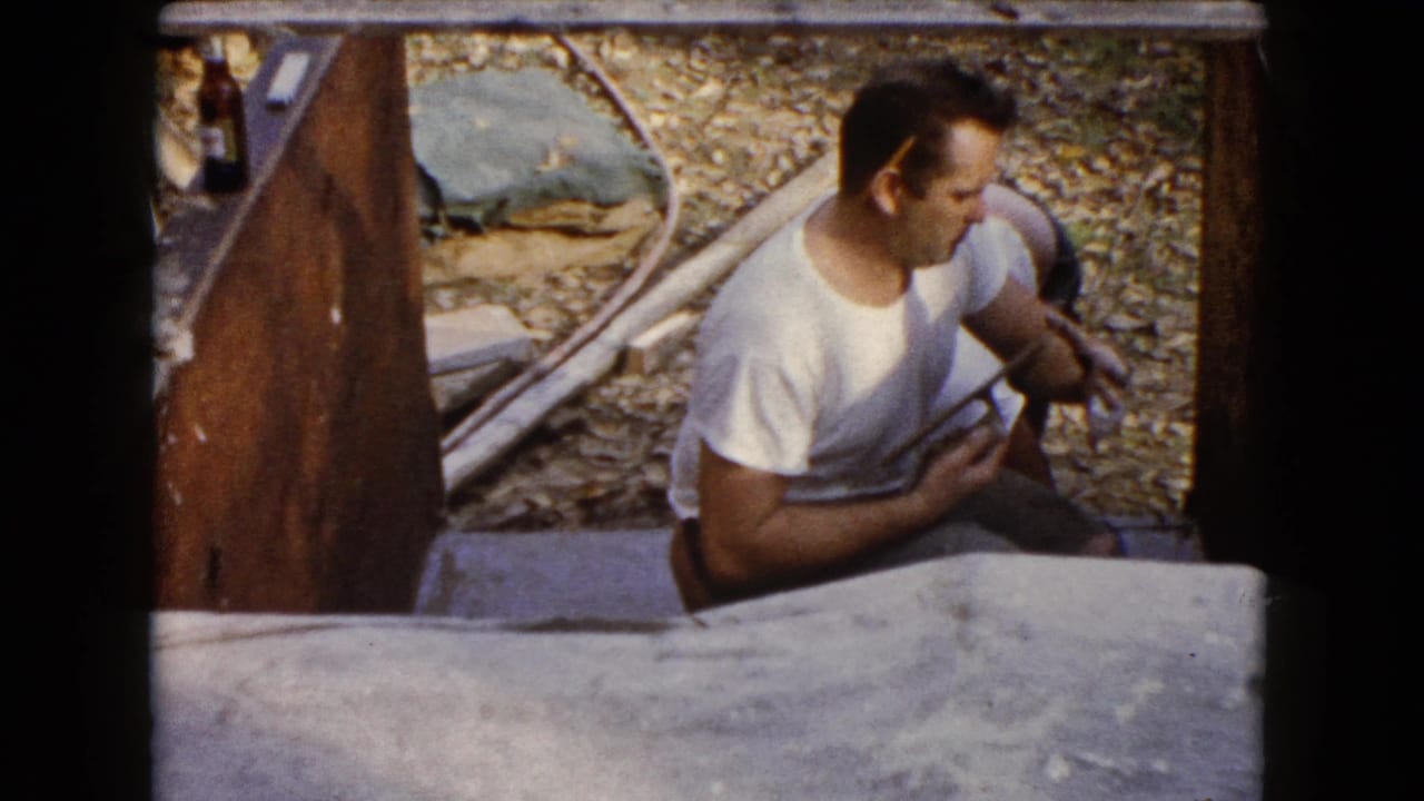The worker turns slightly while holding a tool beside the concrete form, with gravel and boards visible around him.