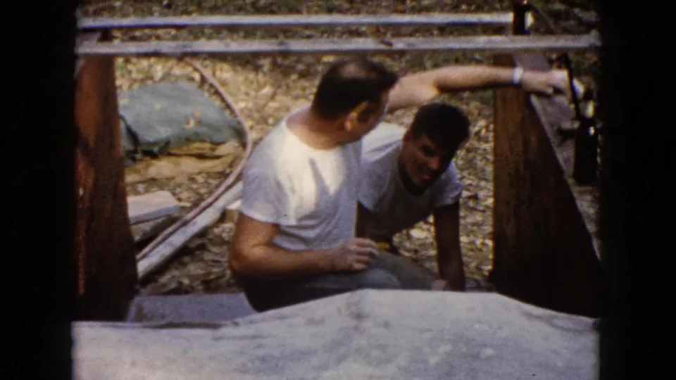 Two men appear in the Wauconda home-movie frame as one reaches toward the wooden form beside the concrete surface.