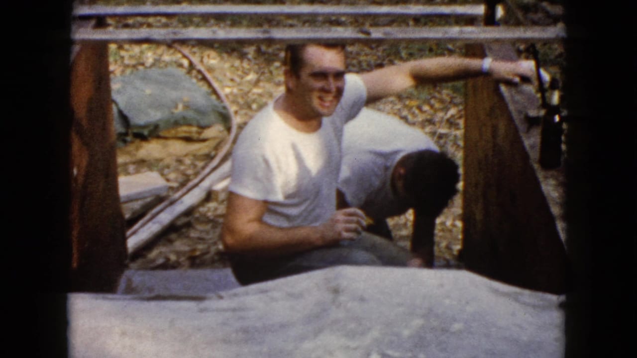 Man in a white T-shirt crouches beside a wooden structure while another man bends nearby.