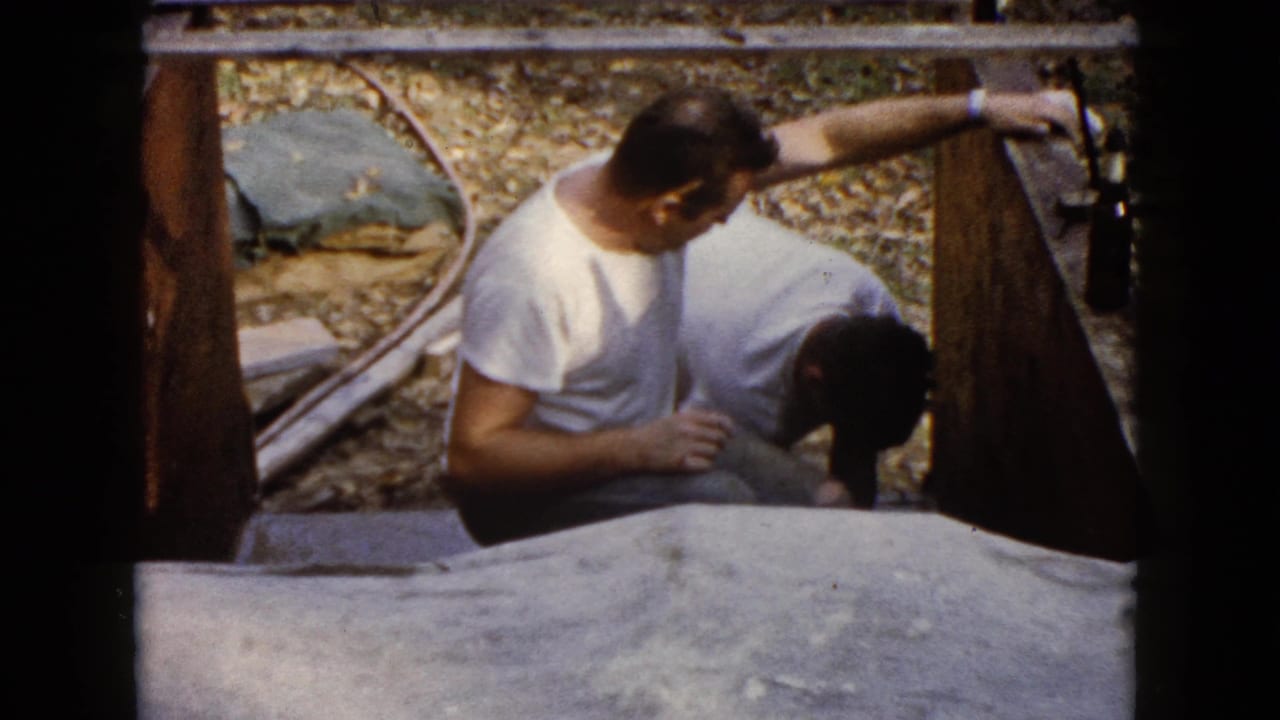 1960 Wauconda frame with a crouching man in a white T-shirt looking down as another man works close to a wooden structure.