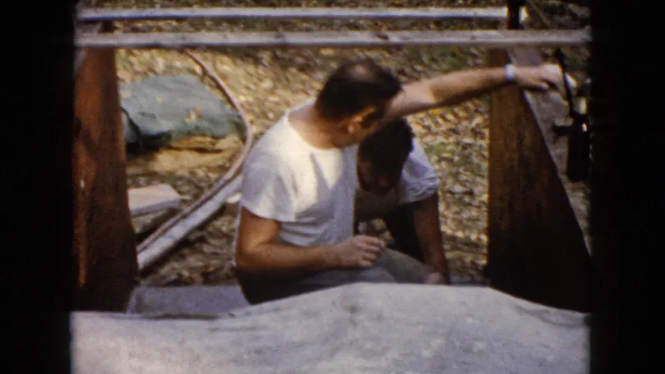 Home-movie frame from 1960 Wauconda showing two men close together at a wooden outdoor work area.