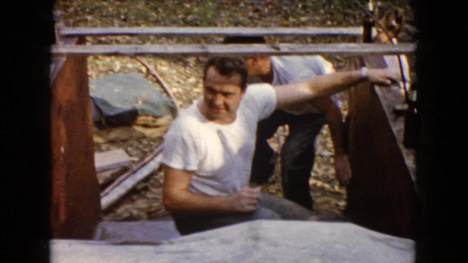 1960 Wauconda 8mm frame showing one man crouched near a large foreground object and another man partly visible behind him.