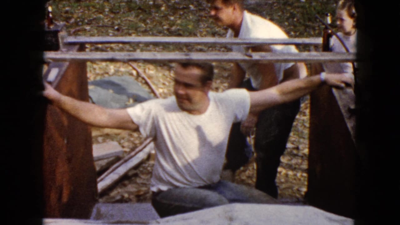 In Wauconda, Illinois, a seated adult man in a white T-shirt leans by a wooden structure as other adults stand in the background.