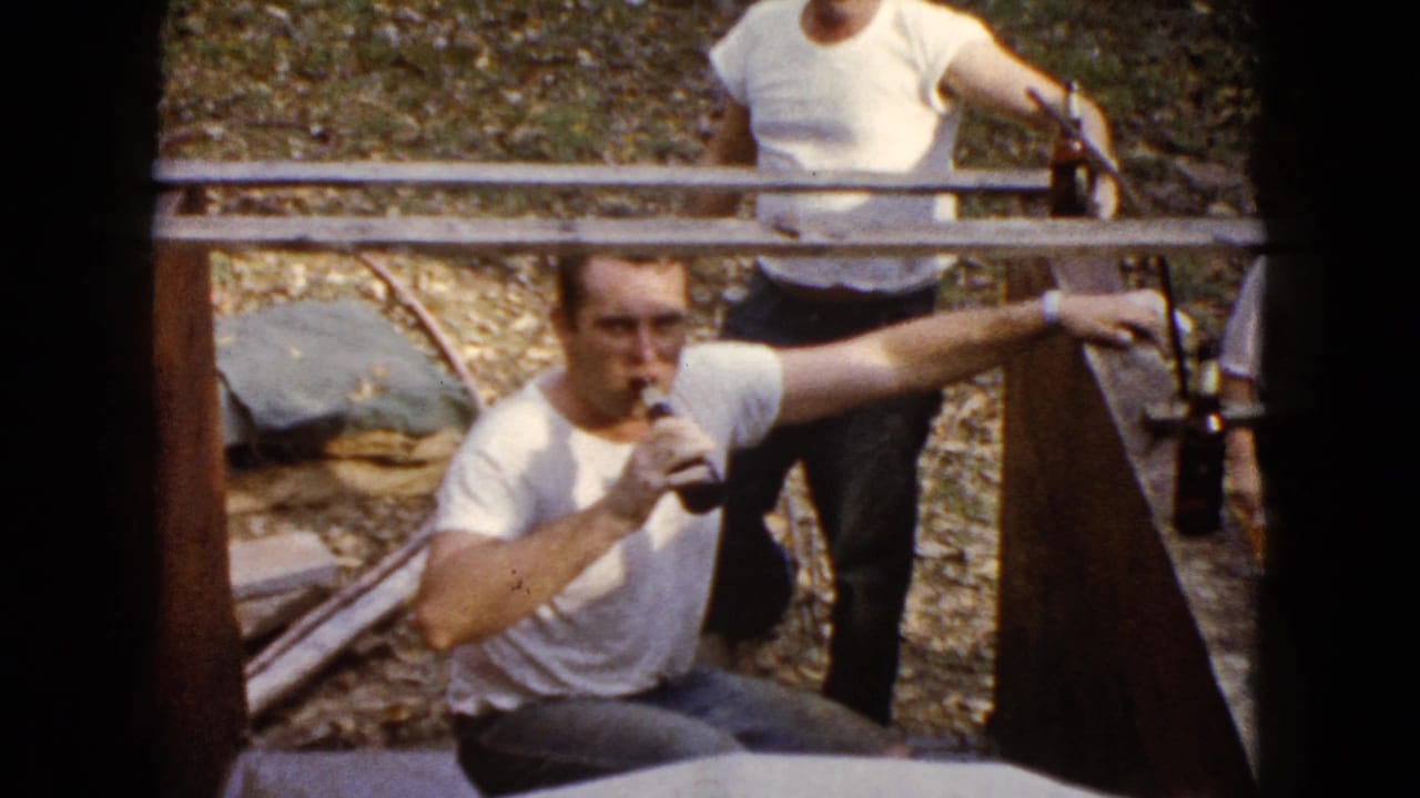 Seated man drinks from a glass bottle while leaning against a wooden structure outdoors.