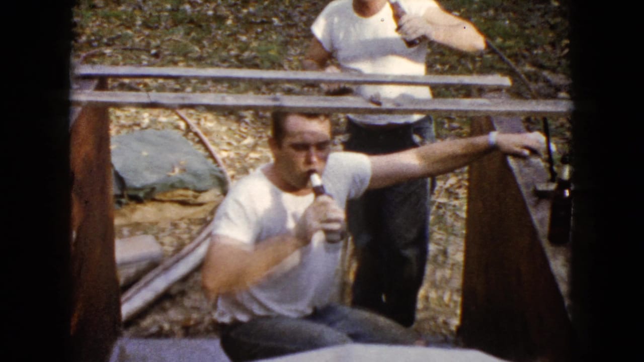 The home-movie frame shows a man in a white T-shirt drinking from a bottle as another adult stands behind him near a rail.