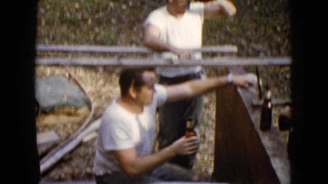 A man in a white T-shirt holds a bottle and turns toward the side near the wooden structure in Wauconda home-movie footage.