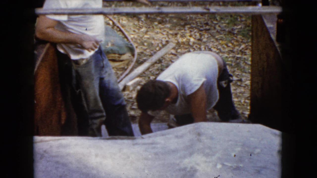 In 1960 Wauconda, Illinois, two adult men in white shirts and jeans work around newly poured concrete steps in an outdoor setting.