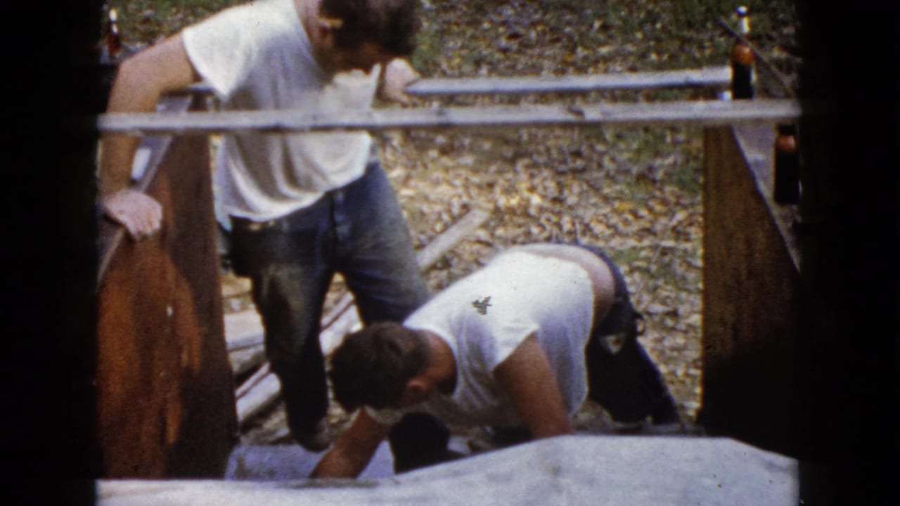 Two adult men in casual work clothes are seen from above as one kneels at the concrete and the other stands beside the wooden form.