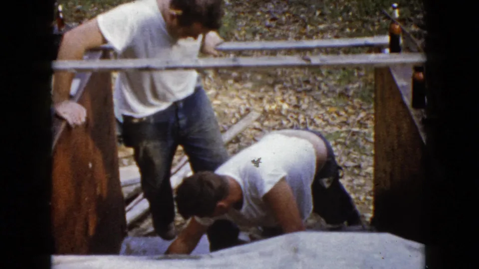 Two adult men in casual work clothes are seen from above as one kneels at the concrete and the other stands beside the wooden form.