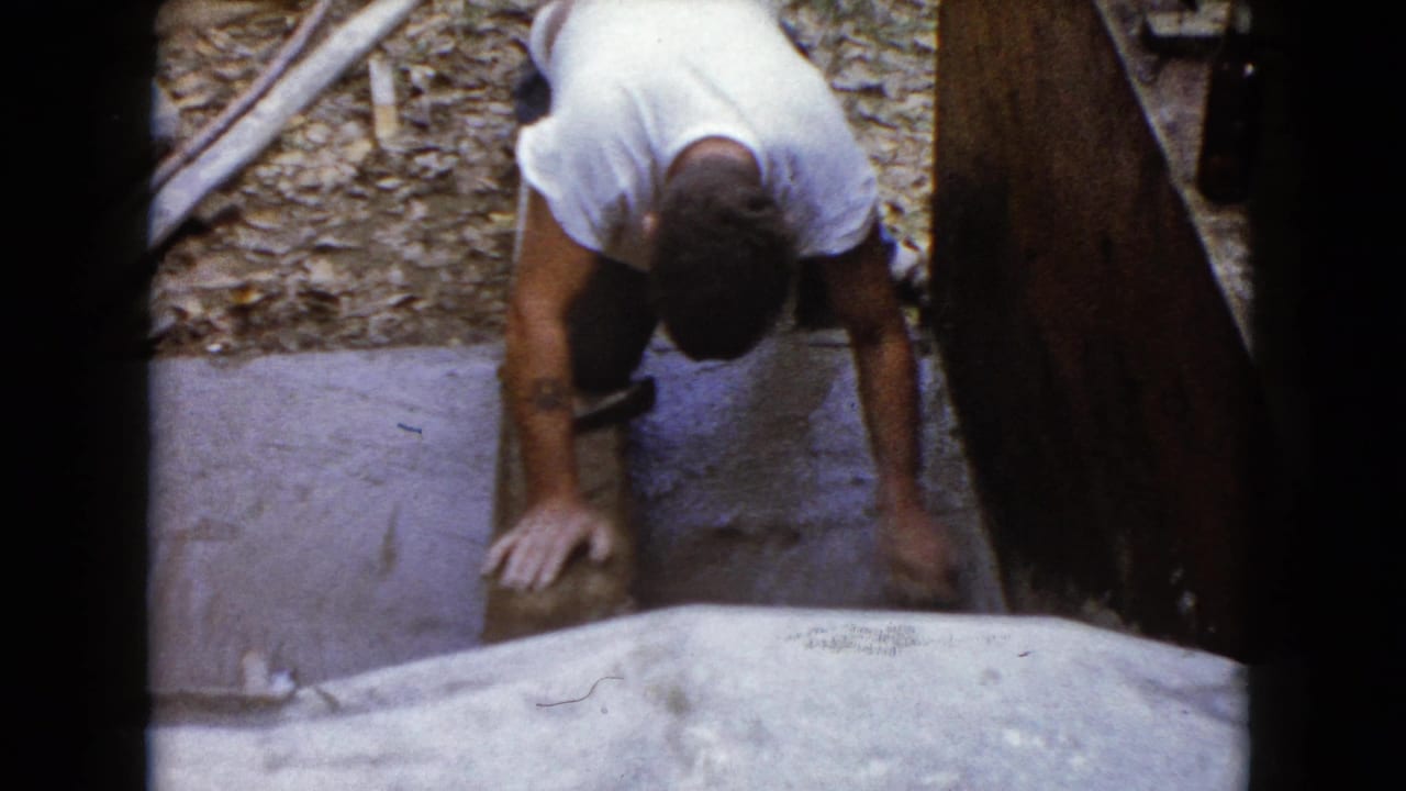 Man kneels outdoors and holds a hammer near a stone or concrete piece.