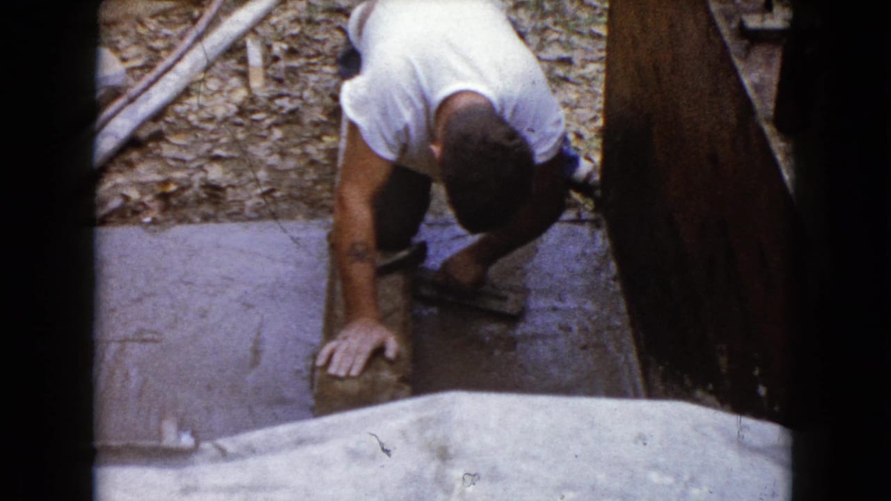 A man in a white T-shirt kneels and leans over a stone or concrete piece while using a hammer in 1960 Wauconda, Illinois home-movie footage.