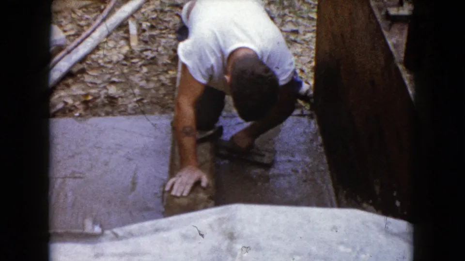 A man in a white T-shirt kneels and leans over a stone or concrete piece while using a hammer in 1960 Wauconda, Illinois home-movie footage.