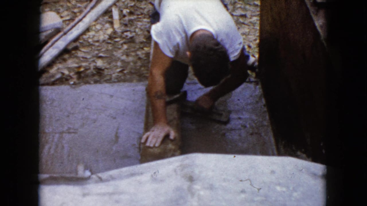 The 8mm frame shows a man crouched at a Wauconda outdoor construction area, bracing one hand while working a hammer near a block.
