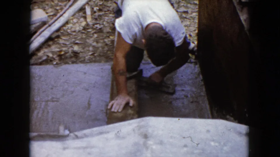 The 8mm frame shows a man crouched at a Wauconda outdoor construction area, bracing one hand while working a hammer near a block.