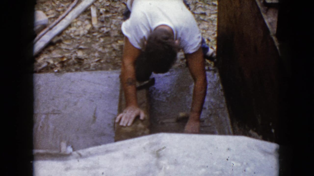 A man kneels near wooden boards and a stone or concrete piece in supported 1960 Wauconda, Illinois 8mm home-movie footage.