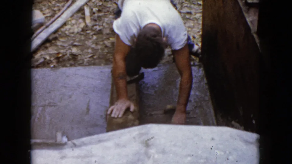 A man kneels near wooden boards and a stone or concrete piece in supported 1960 Wauconda, Illinois 8mm home-movie footage.