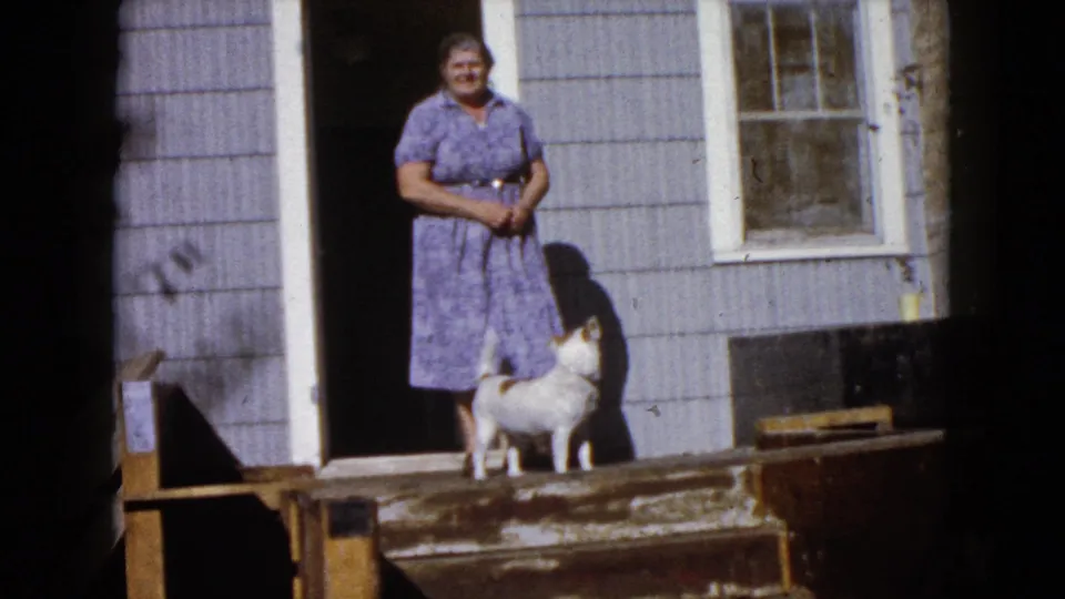 In the 1960 Wauconda home-movie frame, the woman stands facing the camera while the small dog pauses beside her on the stoop.