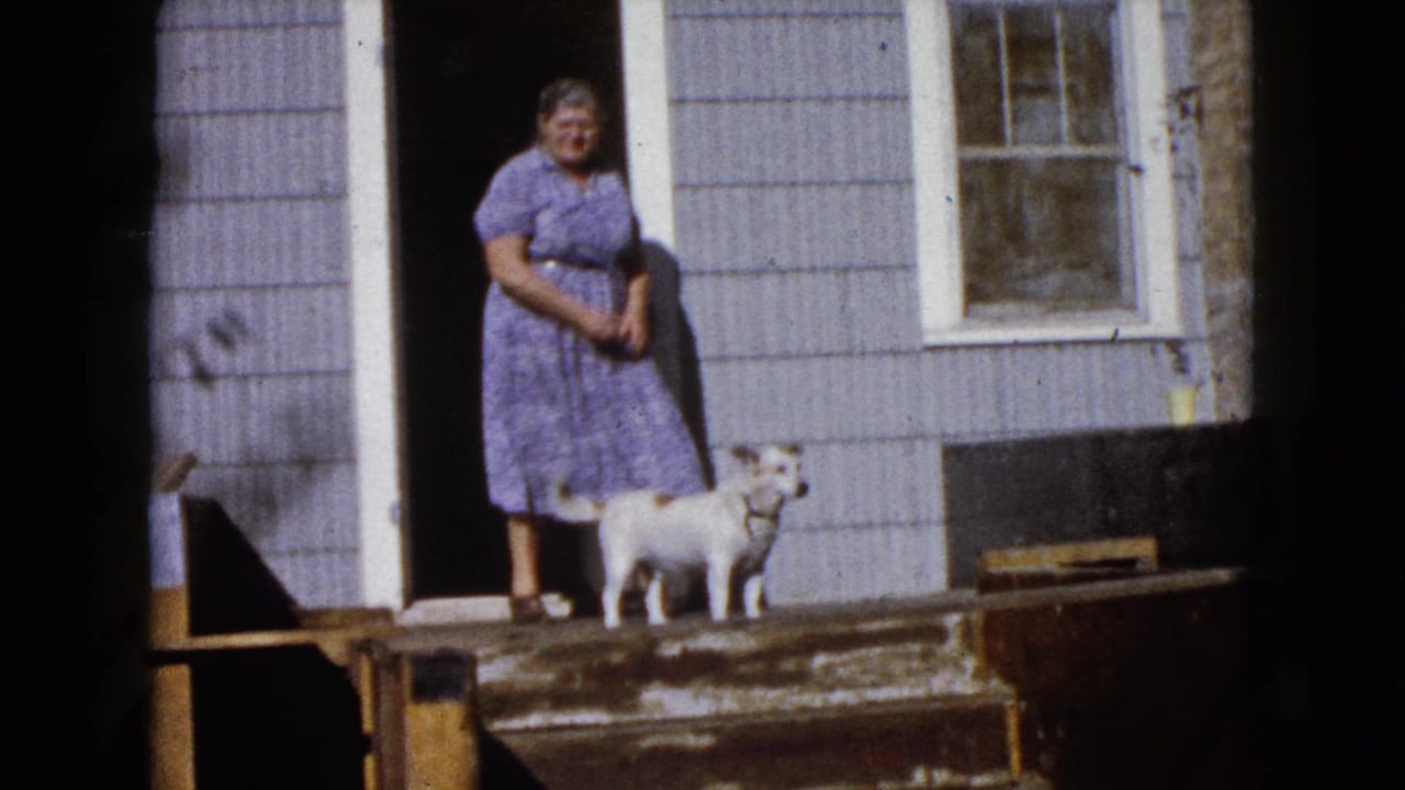 The woman stands in front of the dark doorway of the Wauconda house as the small white dog looks outward from the wooden steps.