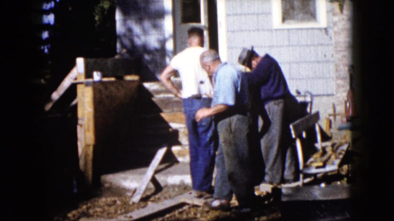 1960 Wauconda frame showing three men gathered beside a wooden structure, hand pump, house siding, and yard steps.